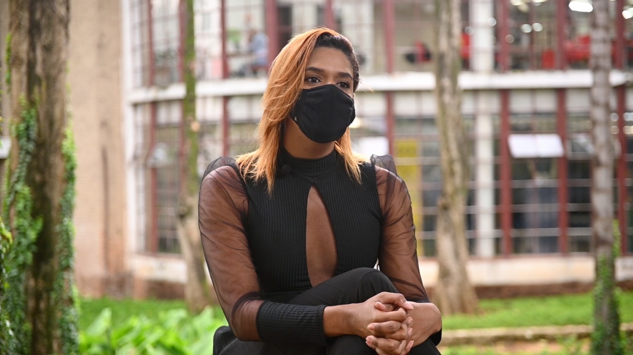 Uma mulher está sentada com as pernas cruzadas e as mãos apoiadas no joelho. Ela veste uma roupa preta, com tom de transparência nos braços e no decote, além de uma máscara também preta. Atrás, está um ambiente aberto, com grama e árvores.